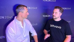 Zwei Männer sitzen und unterhalten sich vor dem Hintergrund einer TOKEN2049-Veranstaltung; einer trägt ein weißes Hemd, der andere ein schwarzes Bitvavo-T-Shirt, während sie über die Strategie für Europas führende Krypto-Börse diskutieren.