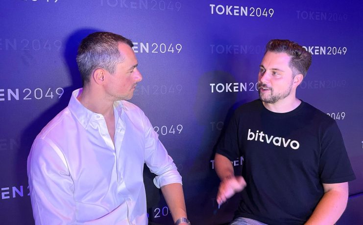 Zwei Männer sitzen und unterhalten sich vor dem Hintergrund einer TOKEN2049-Veranstaltung; einer trägt ein weißes Hemd, der andere ein schwarzes Bitvavo-T-Shirt, während sie über die Strategie für Europas führende Krypto-Börse diskutieren.