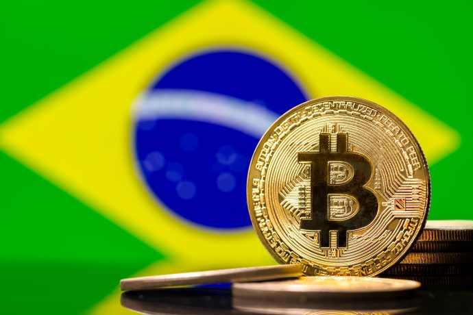 Eine physische Bitcoin-Münze steht vor mehreren Münzen, wobei die brasilianische Flagge im Hintergrund verschwommen ist, was ein wachsendes Interesse an Bitcoin-Strategien in aufstrebenden Fintech-Märkten symbolisiert.