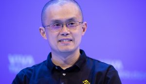 Eine Person mit Brille und einem schwarzen Binance-Poloshirt lächelt vor einem schlichten blauen Hintergrund und erinnert an den ikonischen Look von Changpeng Zhao.