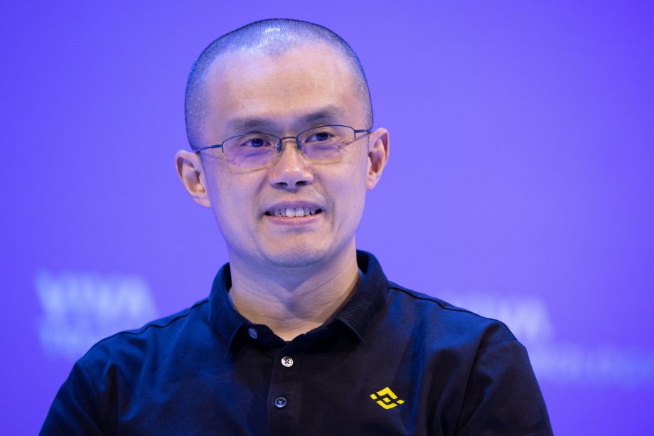 Eine Person mit Brille und einem schwarzen Binance-Poloshirt lächelt vor einem schlichten blauen Hintergrund und erinnert an den ikonischen Look von Changpeng Zhao.