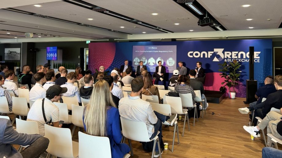 Das Publikum sitzt und hört einer Podiumsdiskussion bei einer CONF3RENCE-Veranstaltung zu, mit Sprechern auf der Bühne und einer Präsentationsleinwand hinter ihnen.