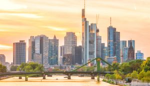 Eine Skyline mit modernen Wolkenkratzern – Sitz führender Krypto-Finanzunternehmen – und einer grünen Brücke über einen Fluss bei Sonnenuntergang, mit Bäumen am Flussufer.