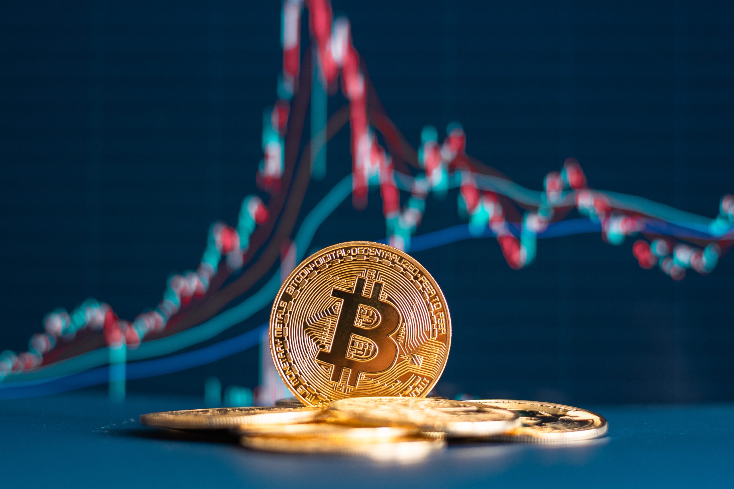 Mehrere physische Bitcoin-Token sind vor einem Bildschirm gestapelt, auf dem ein schwankendes Krypto-Preisdiagramm angezeigt wird.