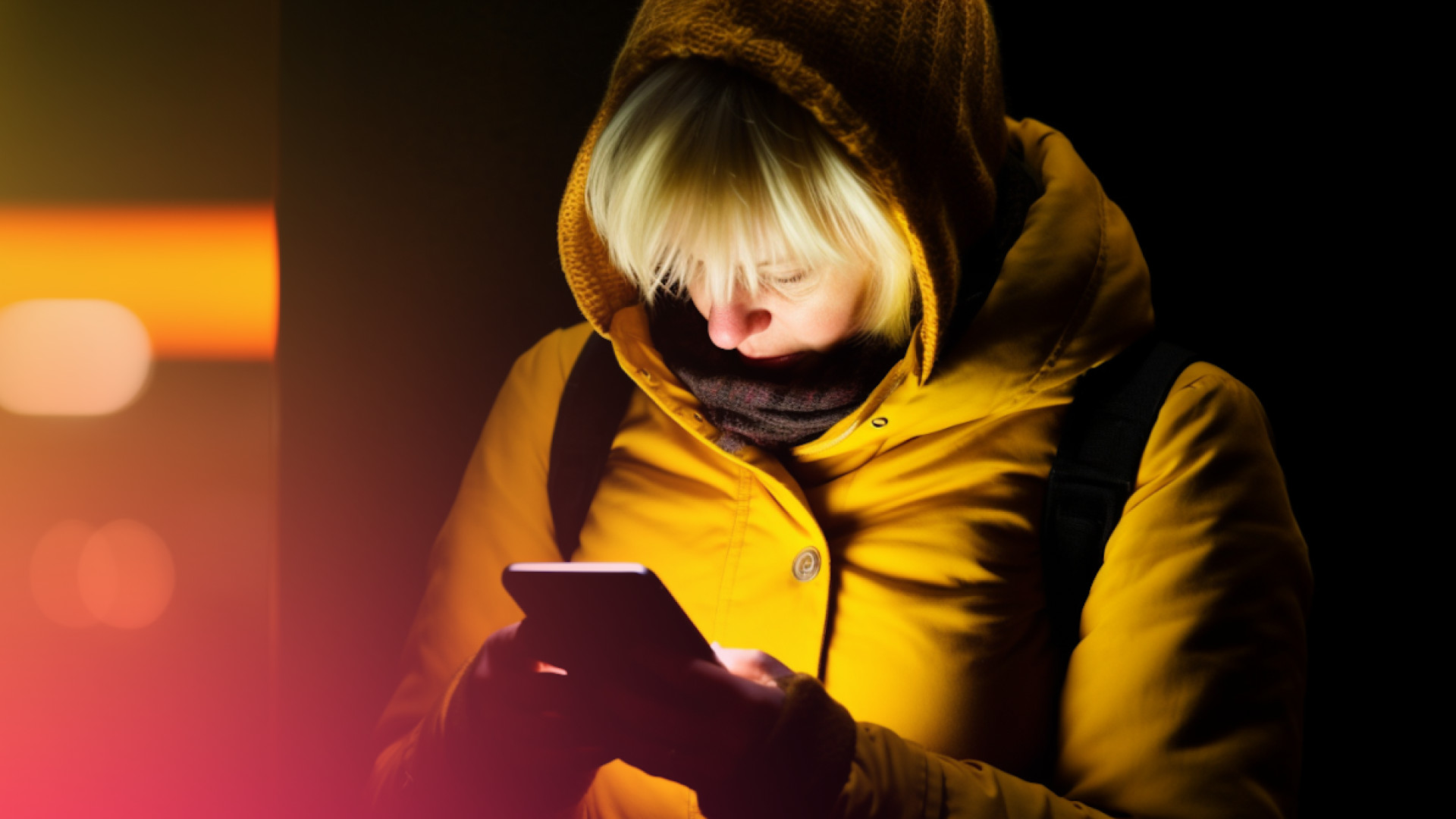 Eine Person, die einen gelb gepunkteten Mantel und eine Kapuze trägt, blickt in einer schwach beleuchteten Umgebung auf ein Smartphone.