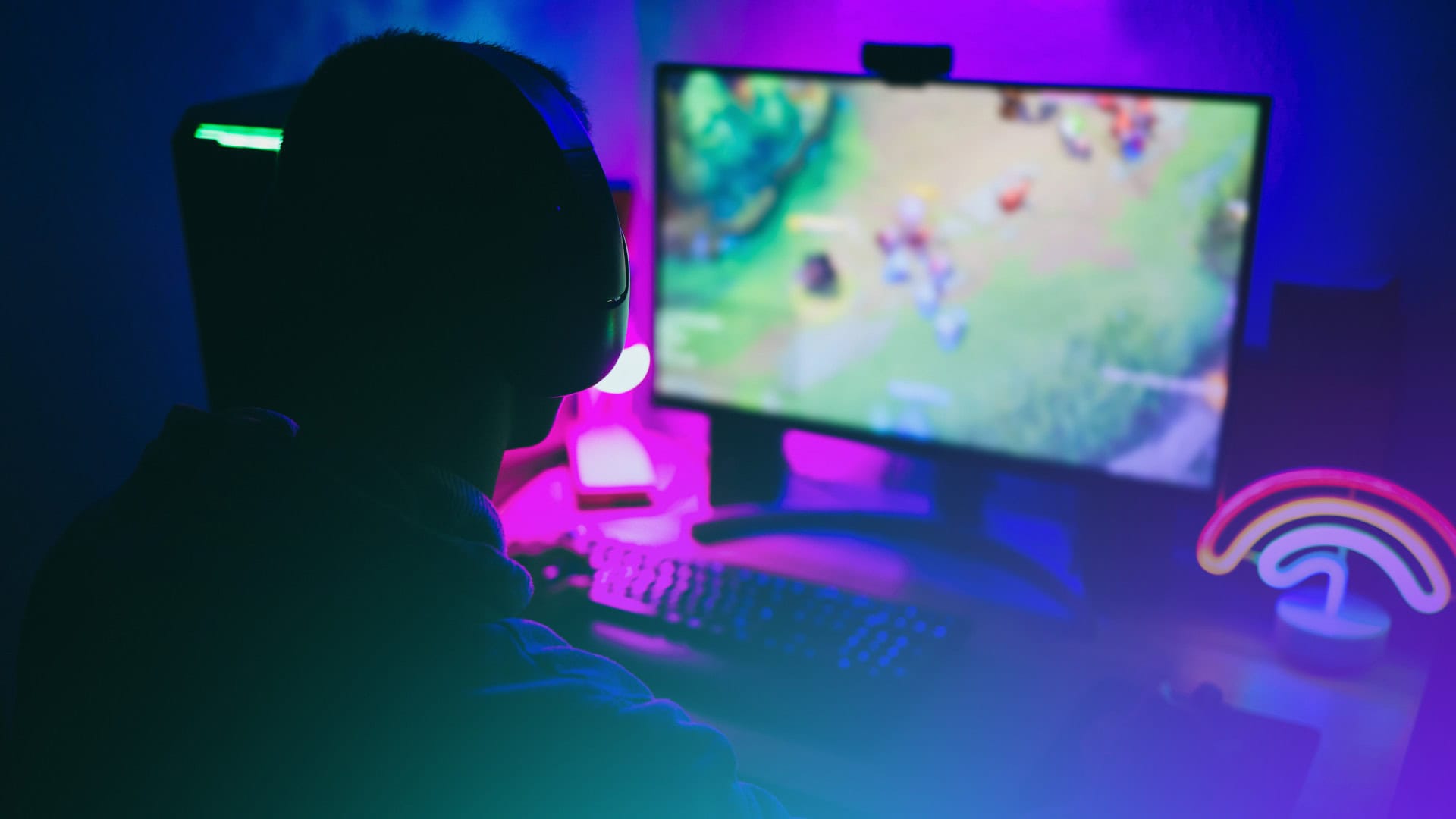 Eine Person mit Kopfhörern sitzt an einem Computertisch und spielt auf einem Monitor in einem schwach beleuchteten Raum mit blauer und violetter Beleuchtung ein farbenfrohes Videospiel mit Polkadot-Thema.
