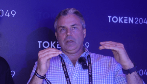 Ein Mann im karierten Hemd spricht mit erhobenen Händen bei TOKEN2049. Er trägt ein Schlüsselband und steht vor einem violetten Hintergrund mit Event-Branding und diskutiert Themen wie Bitcoin.