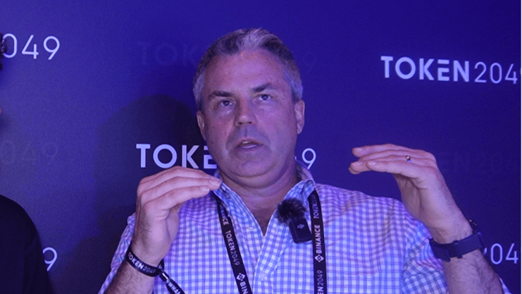 Ein Mann im karierten Hemd spricht mit erhobenen Händen bei TOKEN2049. Er trägt ein Schlüsselband und steht vor einem violetten Hintergrund mit Event-Branding und diskutiert Themen wie Bitcoin.