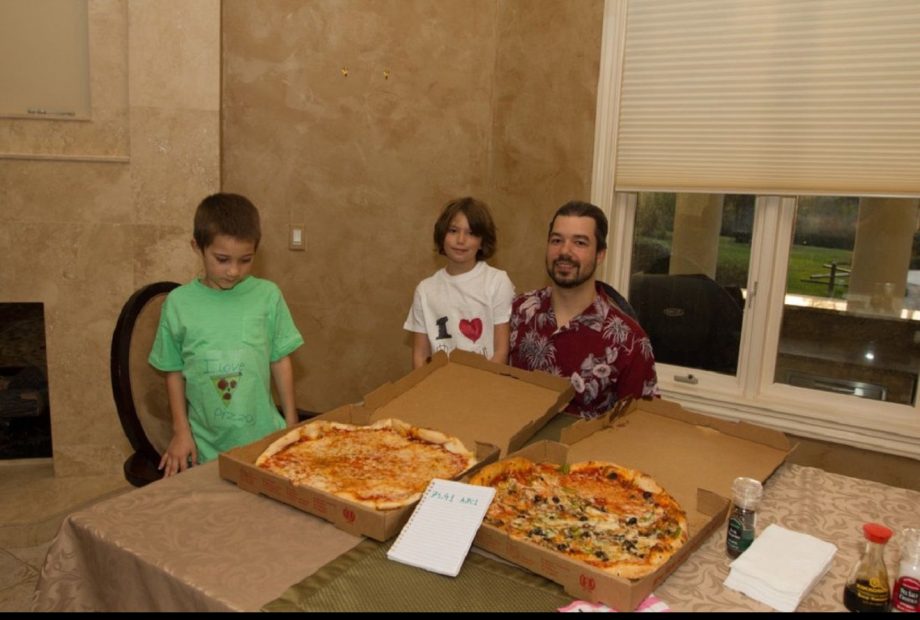 Drei Personen sitzen mit zwei großen Pizzen, einem Notizblock mit Bitcoin-Scheinen und Gewürzen an einem Esstisch in einem Raum mit beigen Wänden und einem Fenster im Hintergrund.