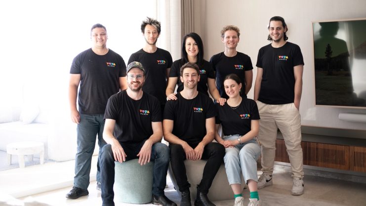 Acht Personen in passenden schwarzen VOYA-T-Shirts mit einem bunten Logo posieren gemeinsam in einem gut beleuchteten, modernen Wohnzimmer, einige stehen, andere sitzen.