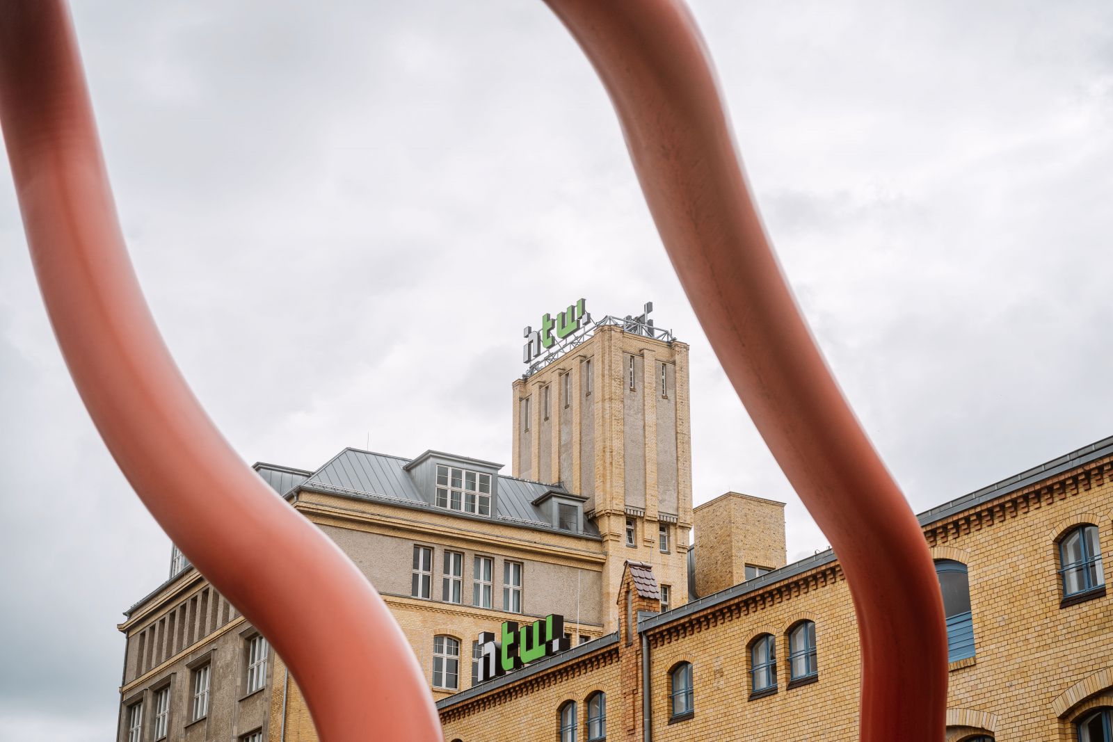 Ein hellbraunes Backsteingebäude mit einem hohen Turm und grünen NTU-Schildern, eingerahmt von zwei geschwungenen roten Metallstrukturen, unter einem bewölkten Himmel.