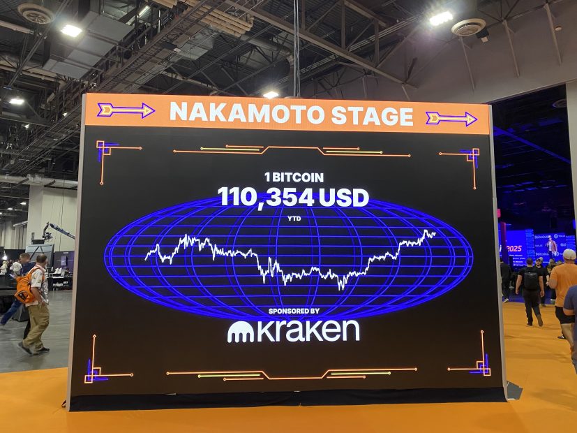 Eine große digitale Anzeige auf der Nakamoto Stage zeigt den Bitcoin-Preis von 110.354 USD neben einem Preisdiagramm und dem Kraken-Logo bei einer Indoor-Veranstaltung mit Menschen im Hintergrund.