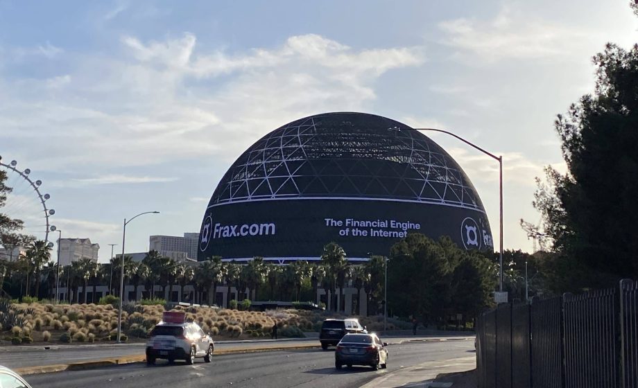 Auf einem großen kugelförmigen Gebäude in Las Vegas ist eine digitale Werbung für Frax.com zu sehen, auf der „The Financial Engine of the Internet“ steht. Im Vordergrund fahren Autos vorbei, als würden sie an einem automatisch gespeicherten, zum Leben erweckten Entwurf vorbeifahren.
