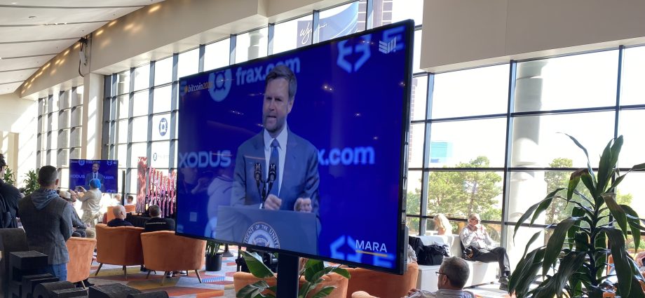 Auf einem Fernsehbildschirm ist ein Mann zu sehen, der während einer Konferenz an einem Podium über Bitcoin spricht. Im Hintergrund sind eine Lounge und große Fenster zu sehen.