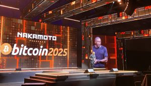 Ein Redner steht auf der Bühne einer Veranstaltung mit großen Bildschirmen, auf denen „NAKAMOTO präsentiert Bitcoin 2025“ angezeigt wird, und diskutiert in einem modernen, beleuchteten Veranstaltungsort den Weg zu 1 Million US-Dollar.