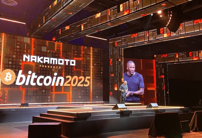 Ein Redner steht auf der Bühne einer Veranstaltung mit großen Bildschirmen, auf denen „NAKAMOTO präsentiert Bitcoin 2025“ angezeigt wird, und diskutiert in einem modernen, beleuchteten Veranstaltungsort den Weg zu 1 Million US-Dollar.