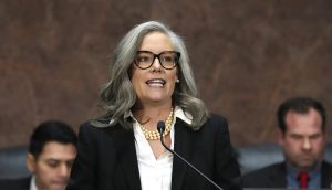 Eine Frau mit grauem Haar und Brille, die einen schwarzen Anzug und eine Perlenkette trägt, spricht an einem Podium in New Hampshire; im Hintergrund sitzen zwei Männer.