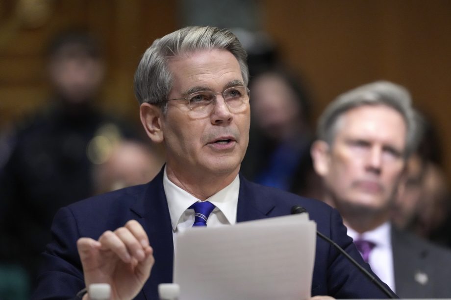 Ein Mann in Anzug und Brille spricht mit Papieren in der Hand bei einem scheinbar formellen Treffen oder einer Anhörung der USA über, während ein anderer Mann im Hintergrund sitzt.