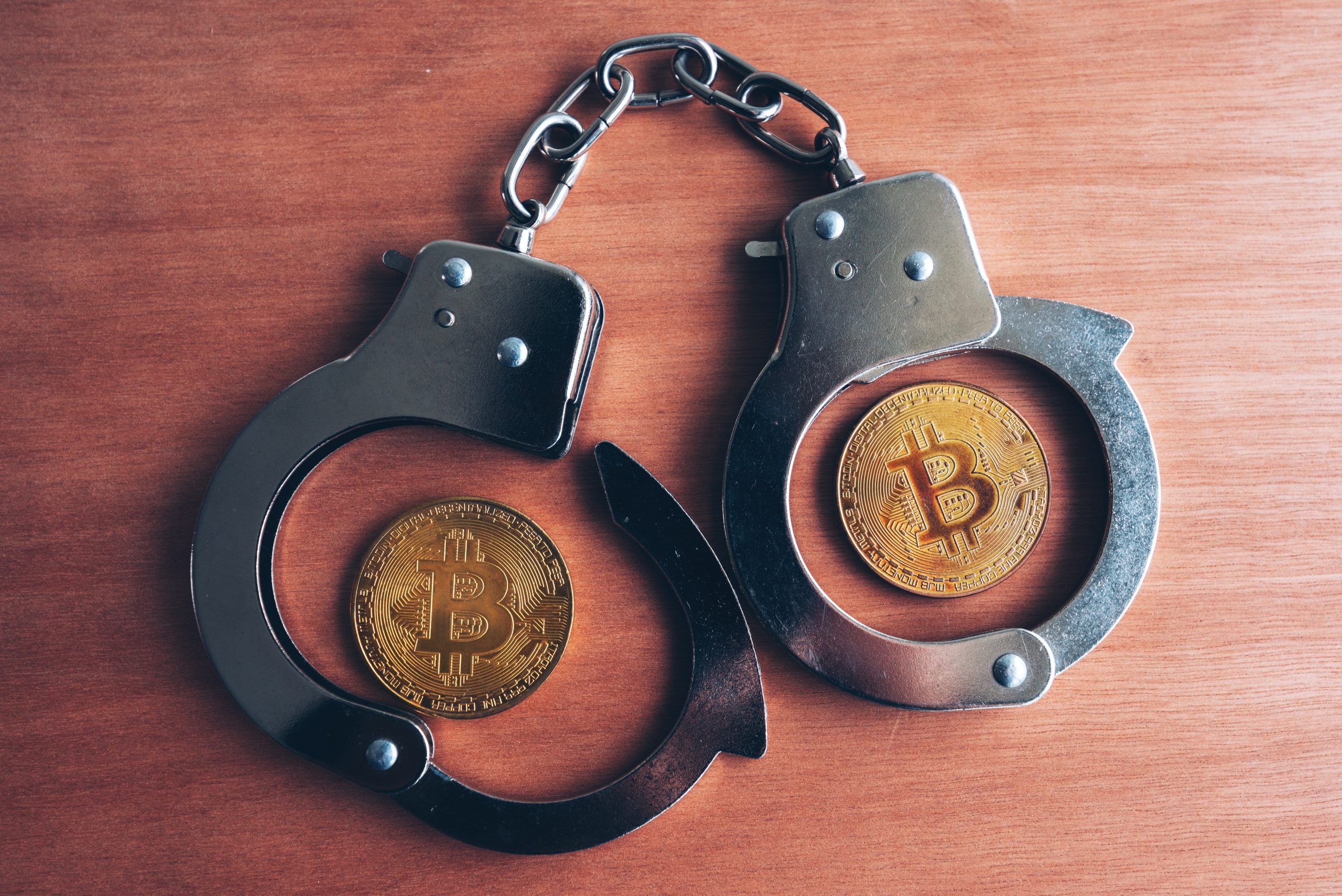 Zwei Bitcoin-Münzen in offenen Handschellen auf einer Holzoberfläche, die Themen wie die Sicherheit von Krypto-Millionären und die potenziellen Risiken von Entführungen in der Welt der Kryptowährungen und der Regulierung aufgreifen.