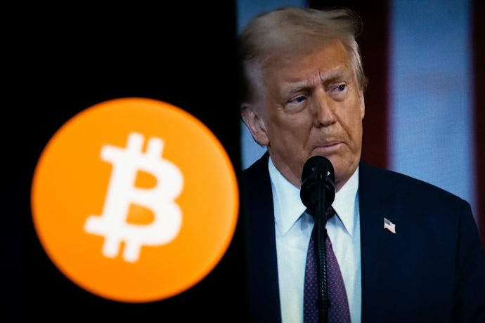 Ein Mann im Anzug spricht in ein Mikrofon, das teilweise von einem großen orangefarbenen Bitcoin-Logo im Vordergrund verdeckt wird und die neuesten Nachrichten vom Krypto-Markt hervorhebt.