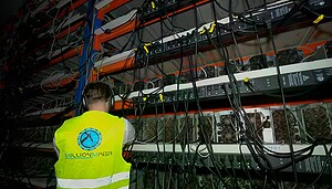 Eine Person in einer gut sichtbaren Weste mit der Aufschrift MillionMiner überwacht mehrere Kryptowährungs-Mining-Maschinen, die auf Regalen angeordnet sind.