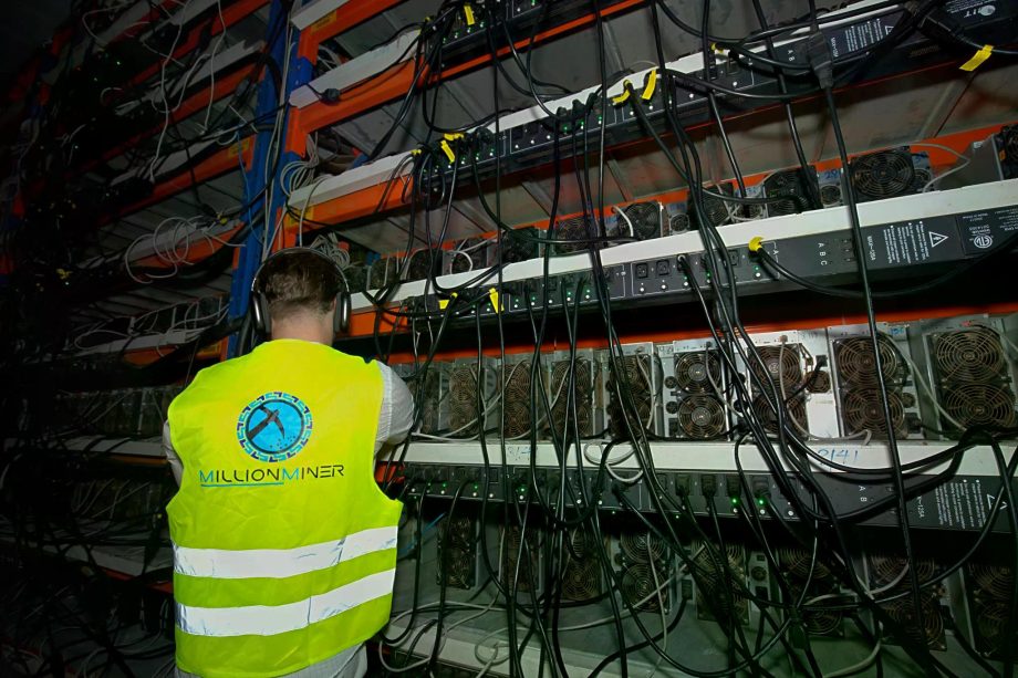 Eine Person in einer gut sichtbaren Weste mit der Aufschrift MillionMiner überwacht mehrere Kryptowährungs-Mining-Maschinen, die auf Regalen angeordnet sind.
