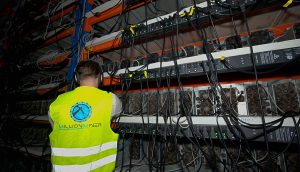 Eine Person mit einer Warnweste steht vor Regalen voller Kryptowährungs-Mining-Maschinen von MillionMiner und zahlreichen Kabeln.