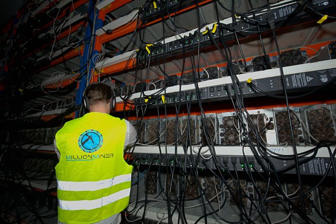 Eine Person mit einer Warnweste steht vor Regalen voller Kryptowährungs-Mining-Maschinen von MillionMiner und zahlreichen Kabeln.