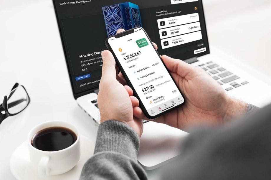 Eine Person hält ein Smartphone mit der Finanz-App MillionMiner, sitzt an einem Schreibtisch mit einem Laptop, auf dem ein Dashboard angezeigt wird, und neben sich steht eine Tasse Kaffee.