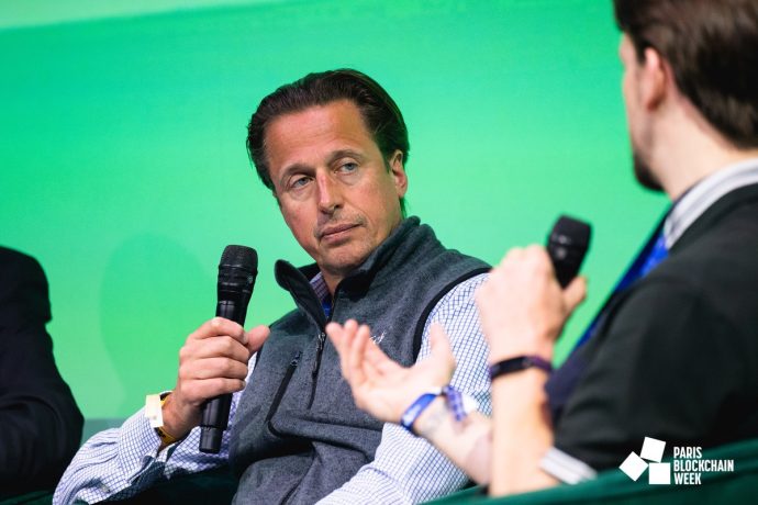 Zwei Männer nehmen an einer Podiumsdiskussion bei der Paris Blockchain Week teil. Einer hält ein Mikrofon und hört aufmerksam zu, während der andere während eines Vortrags mit VanEck mit den Händen gestikuliert.