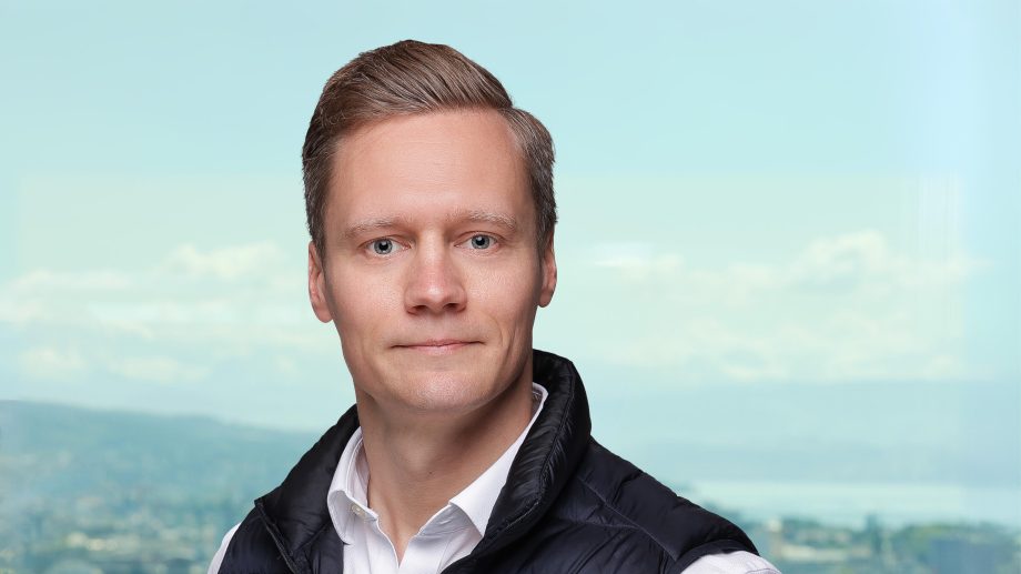 A man with a short, light brown hair, a white shirt and a black vest stands in front of a window with a blurred outdoor landscape and radiates a confidence that can often be seen in the fast-moving world of crypto finances.