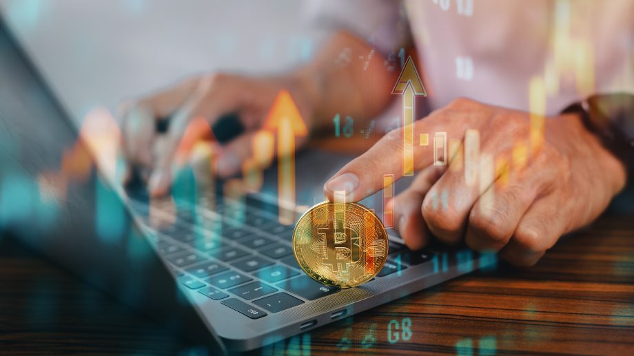 Eine Person tippt auf einem Laptop, im Vordergrund ist eine physische Bitcoin-Münze zu sehen, und aufwärts zeigende Finanzdiagramme symbolisieren den Handel mit Kryptowährungen oder Investitionen mit Revolut für europäische Kunden.
