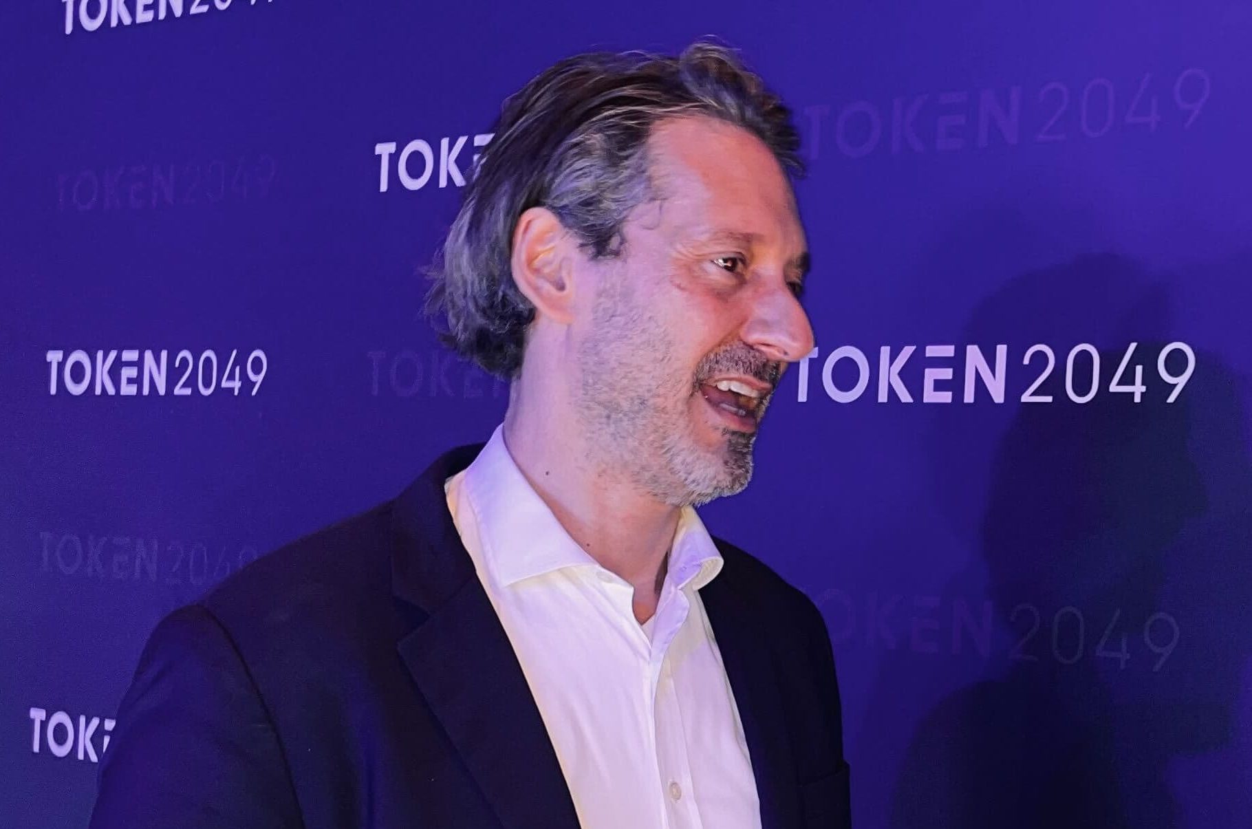 Ein Mann in einem weißen Hemd und einem dunklen Blazer steht vor einem blauen Hintergrund, auf dem wiederholt die Logos von TOKEN2049 angezeigt werden
