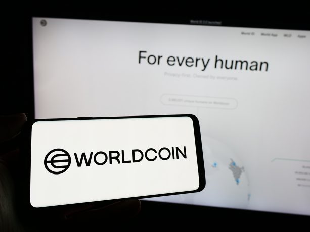 Auf einem Smartphone wird das Worldcoin-Logo vor einem Computerbildschirm angezeigt, auf dem die Worldcoin-Website mit dem Text „Für jeden Menschen“ angezeigt wird, wobei „Weltzugang für alle“ hervorgehoben wird.