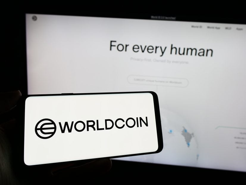 Auf einem Smartphone wird das Worldcoin-Logo vor einem Computerbildschirm angezeigt, auf dem die Worldcoin-Website mit dem Text „Für jeden Menschen“ angezeigt wird, wobei „Weltzugang für alle“ hervorgehoben wird.