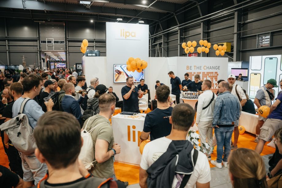 Eine Menschenmenge versammelt sich um einen Stand mit der Aufschrift „Lipa“ im BTC Prag. Die Leute hören einem Redner zu und der Bereich ist mit orangefarbenen Luftballons dekoriert.