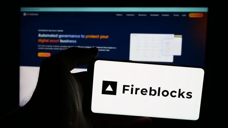 Eine Person hält ein Smartphone mit dem Fireblocks-Logo vor einen Computerbildschirm, auf dem die Homepage der Fireblocks-Website angezeigt wird. Dies verdeutlicht, wie TradFi-Institutionen zunehmend aktiv Stablecoins nutzen.