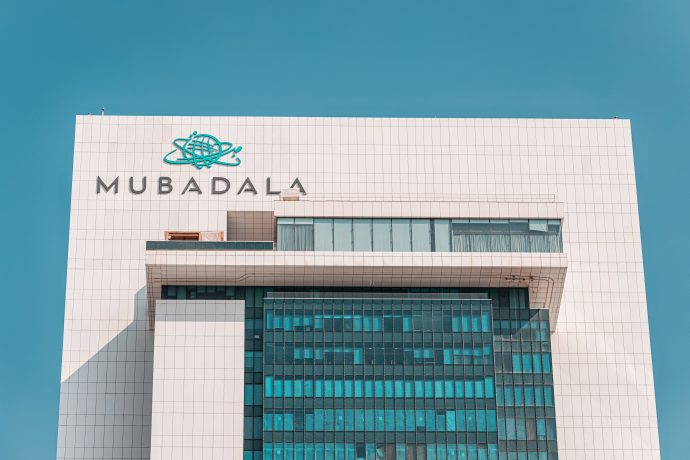 Ein modernes Hochhaus mit dem Wort Mubadala und einem blauen Logo auf der weißen Fassade vor einem klaren blauen Himmel in Abu Dhabi.