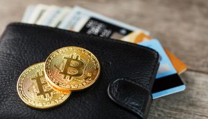 Auf einer Holzoberfläche liegt eine schwarze Brieftasche mit Bargeld und Kreditkarten darin, darauf liegen zwei glänzende Bitcoin-Münzen.