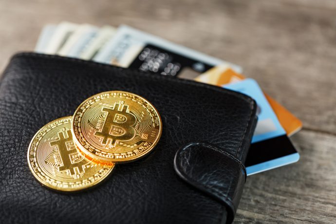 Auf einer Holzoberfläche liegt eine schwarze Brieftasche mit Bargeld und Kreditkarten darin, darauf liegen zwei glänzende Bitcoin-Münzen.