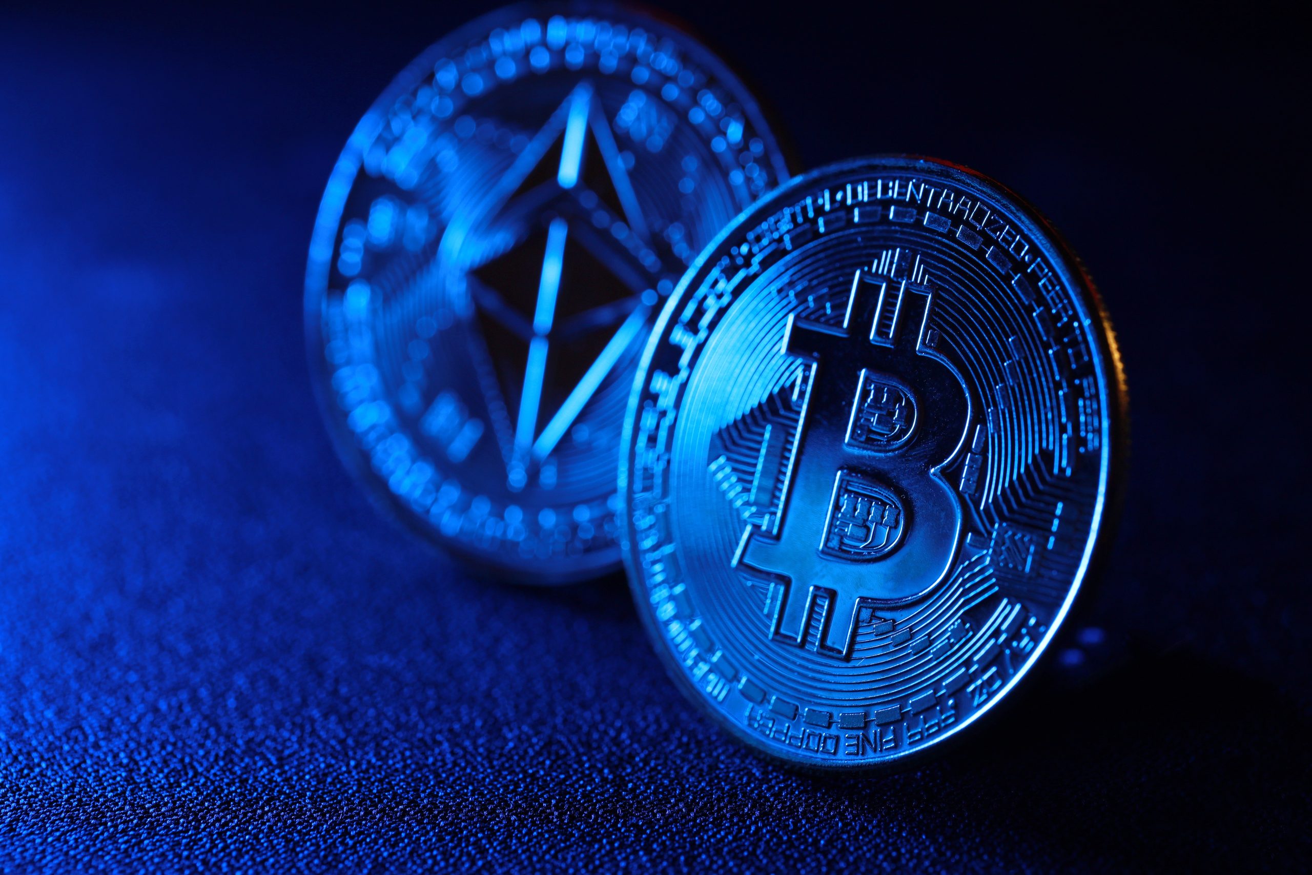 Nahaufnahme physischer Darstellungen von Bitcoin- und Ethereum-Münzen, die aufrecht auf einer dunklen, strukturierten Oberfläche platziert und mit blauem Licht beleuchtet sind – wie ein automatisch gespeicherter Entwurf der digitalen Kryptowelt.