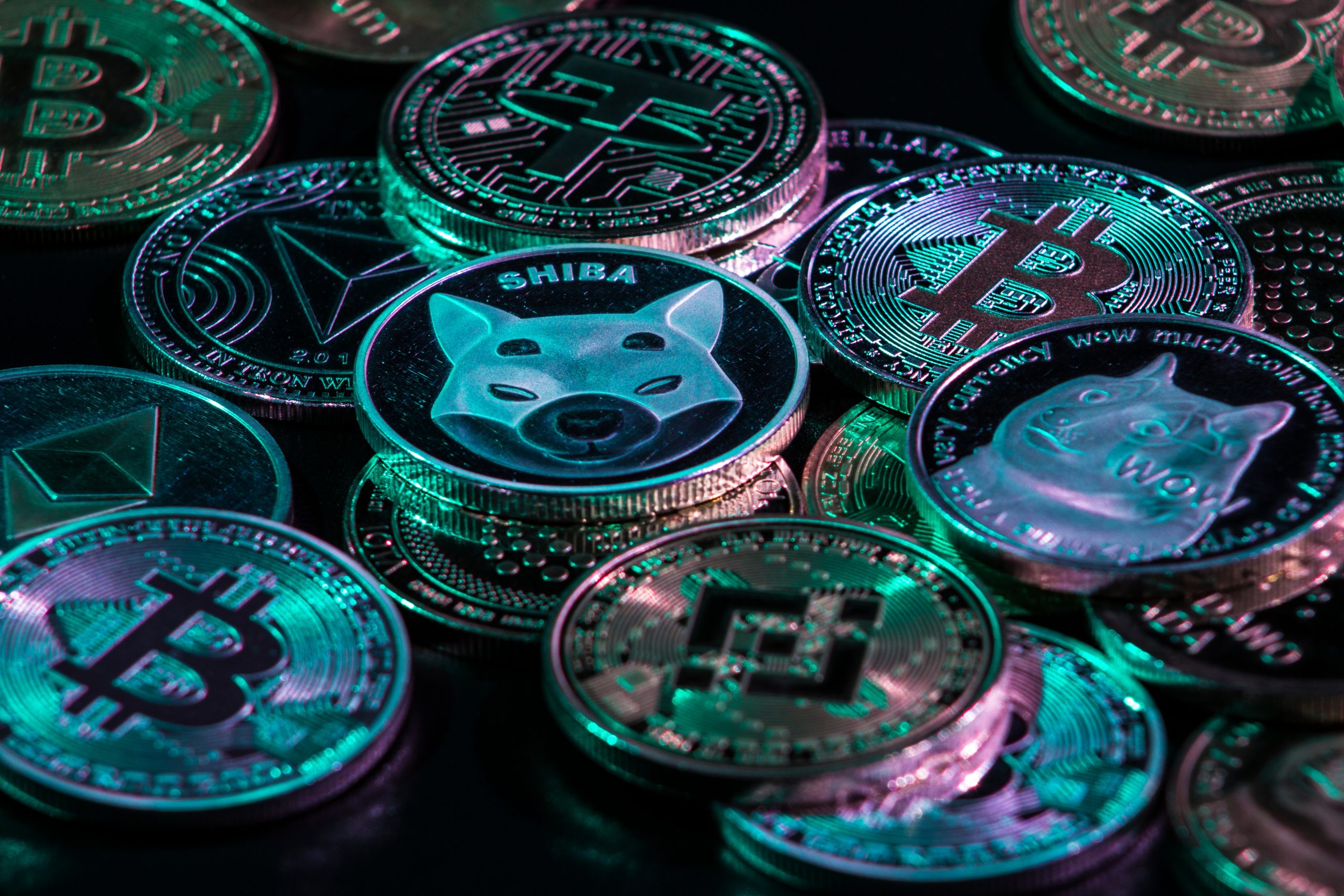 Stapel verschiedener physischer Kryptowährungsmünzen, darunter Bitcoin-, Ethereum-, Binance-, Dogecoin- und Shiba-Inu-Token, beleuchtet mit blauem und grünem Licht.