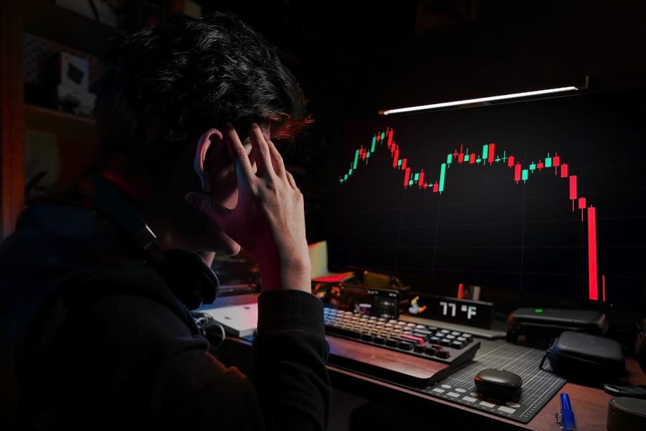 Eine Person sitzt an einem Schreibtisch, hält den Kopf in der Hand und blickt auf einen Computerbildschirm, auf dem ein starker Rückgang in einem Candlestick-Aktienchart angezeigt wird.