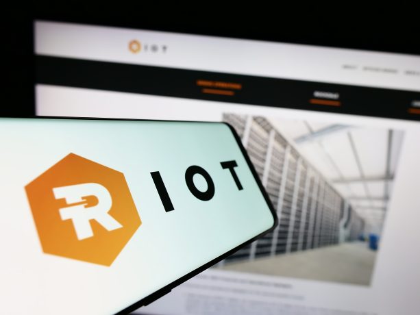 Auf einem Smartphone wird das Logo von Riot Platforms vor einem Computerbildschirm angezeigt, auf dem die Website des Unternehmens mit einem Bild eines Serverraums dargestellt ist.