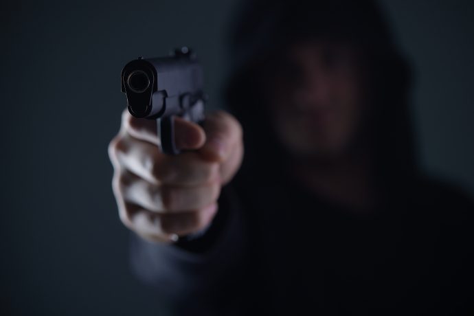 Eine Person mit einem dunklen Kapuzenpullover richtet eine Pistole auf die Kamera, was auf einen Krypto-Raub hindeutet. Die Waffe ist scharf abgebildet, während das Gesicht im Hintergrund unscharf bleibt.