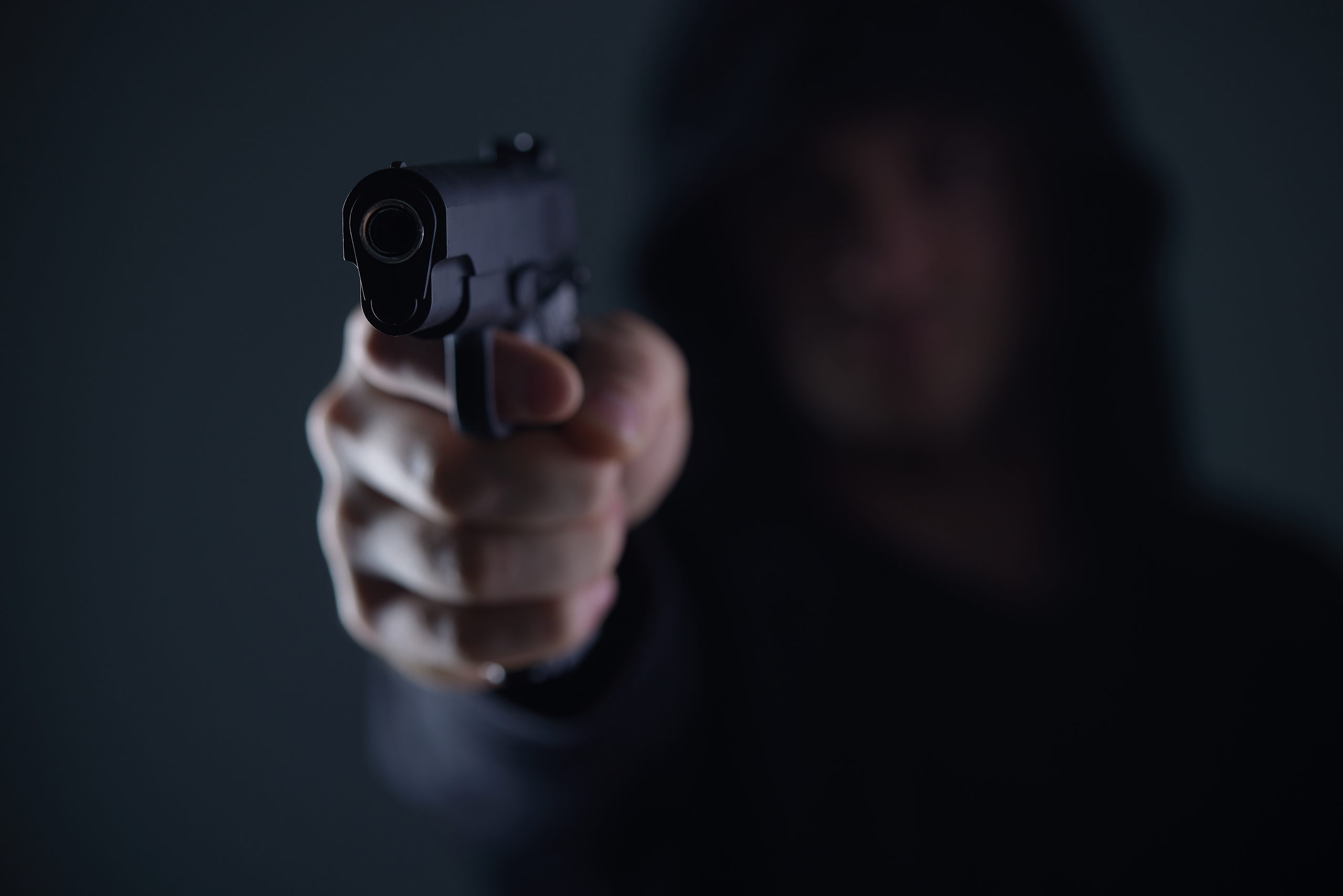 Eine Person mit einem dunklen Kapuzenpullover richtet eine Pistole auf die Kamera, was auf einen Krypto-Raub hindeutet. Die Waffe ist scharf abgebildet, während das Gesicht im Hintergrund unscharf bleibt.