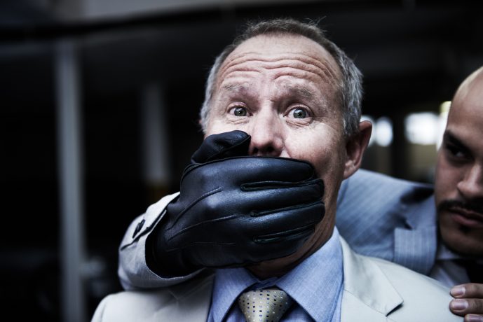 Ein Mann im Anzug hält sich mit einer behandschuhten Hand von hinten den Mund zu, während ein anderer Mann in seiner Nähe steht und ihn aufmerksam beäugt – was der Szene einen Hauch kryptogetriebener Intrige verleiht.