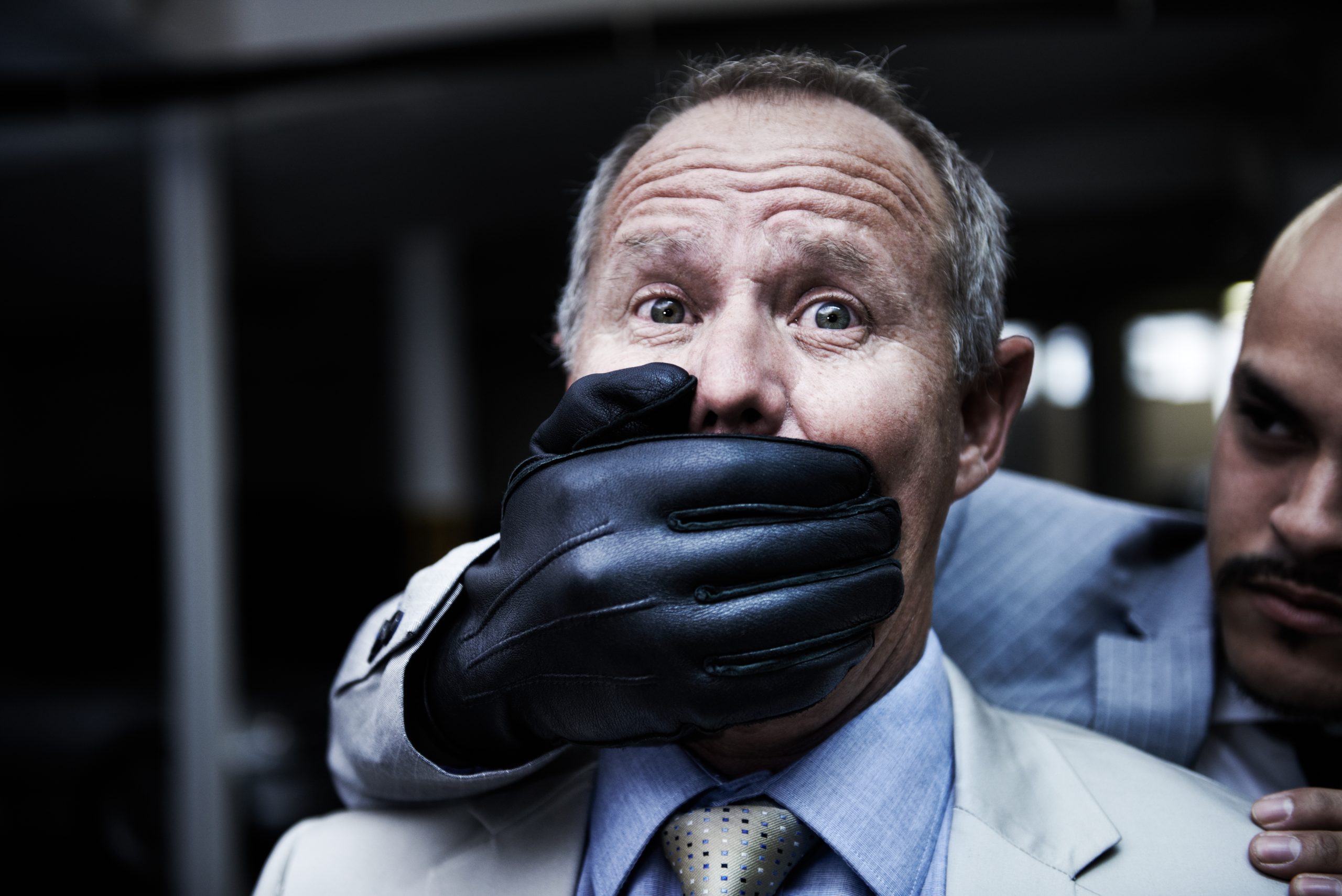 Ein Mann im Anzug hält sich mit einer behandschuhten Hand von hinten den Mund zu, während ein anderer Mann in seiner Nähe steht und ihn aufmerksam beäugt – was der Szene einen Hauch kryptogetriebener Intrige verleiht.