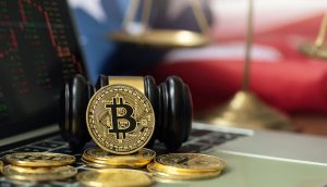 Eine Bitcoin-Münze liegt auf einem Richterhammer, umgeben von Krypto-Münzen. Im Hintergrund sind auf einem Laptop Aktiencharts und eine verschwommene amerikanische Flagge und Waagen zu sehen.