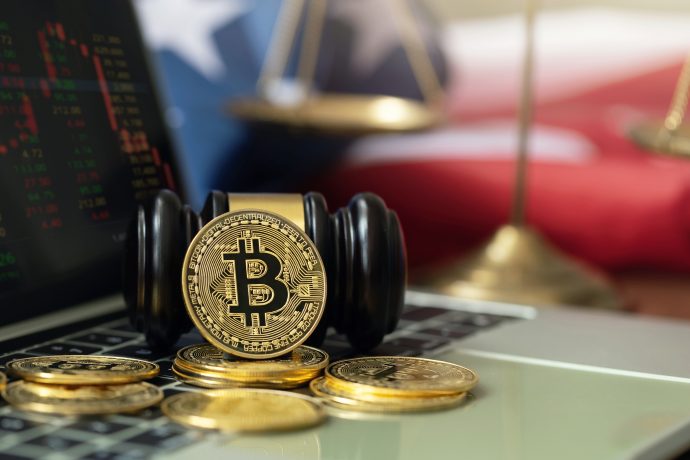 Eine Bitcoin-Münze liegt auf einem Richterhammer, umgeben von Krypto-Münzen. Im Hintergrund sind auf einem Laptop Aktiencharts und eine verschwommene amerikanische Flagge und Waagen zu sehen.